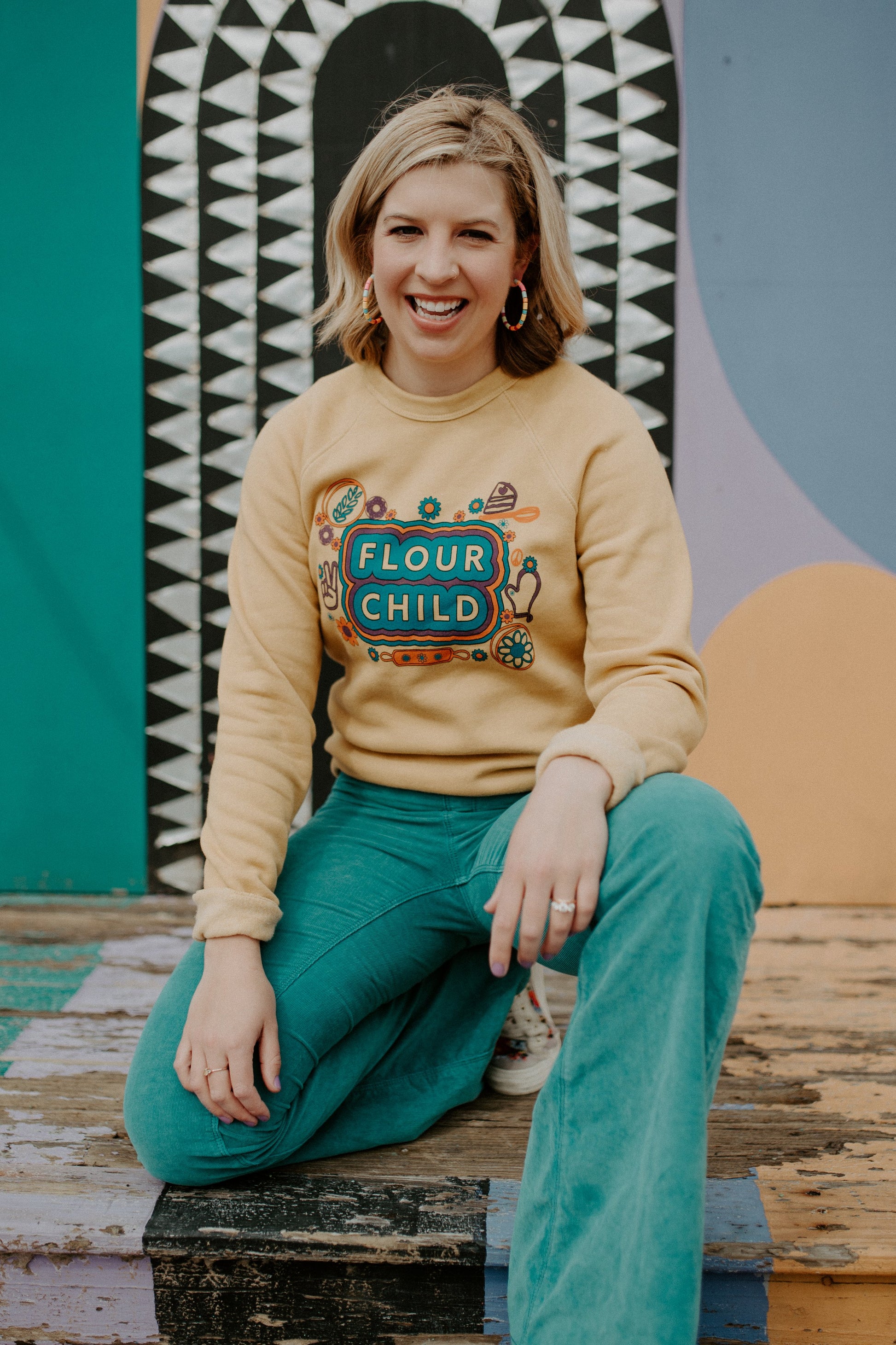 A woman in a bright yellow Flour Child sweatshirt and green pants laughing in front of a mural