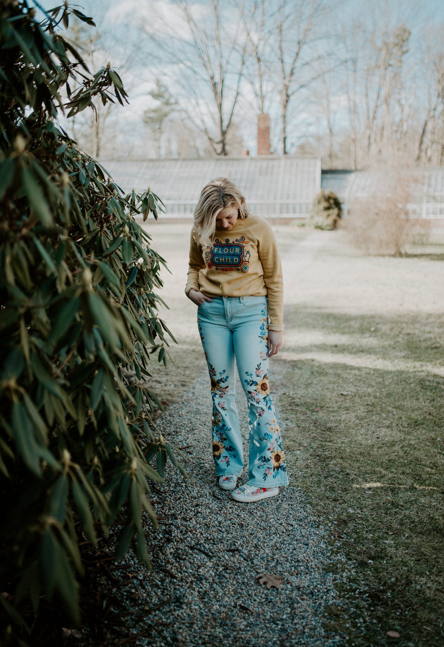 A woman standing outside wears a yellow Flour Child crewneck and light embroidered jeans