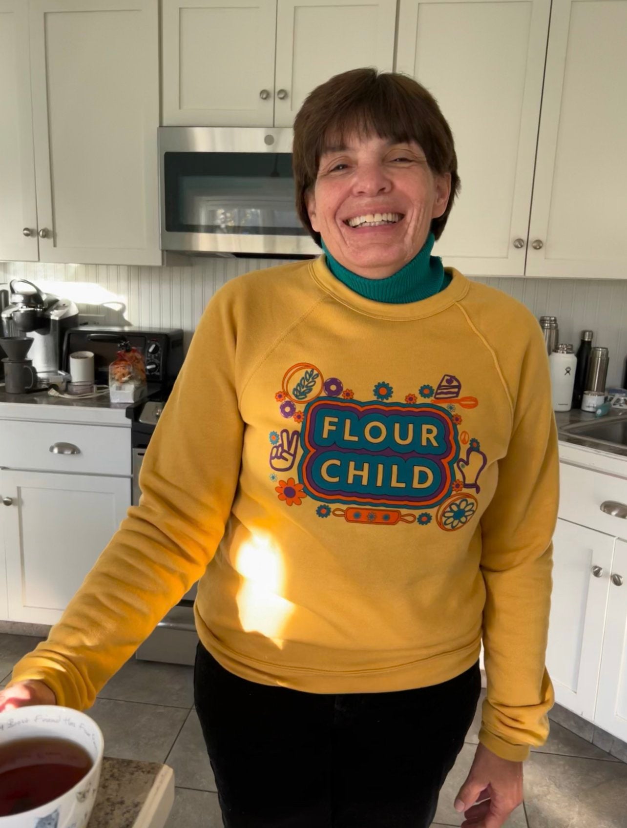 A woman holding a cup of tea smiles wearing the yellow Flour Child crewneck over a green turtleneck