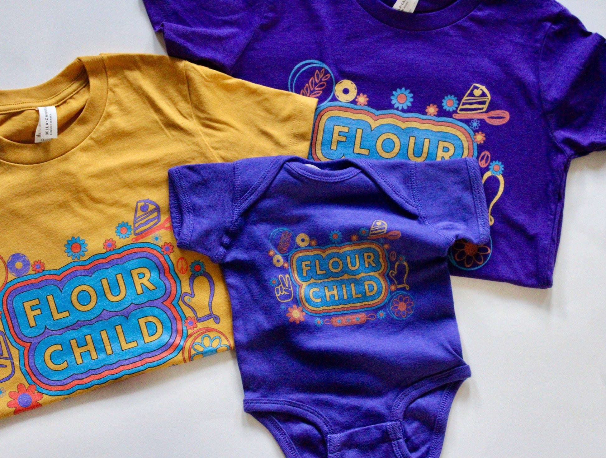 A yellow youth tee, a purple youth tee and a purple baby onesie with the words Flour Child and colorful designs