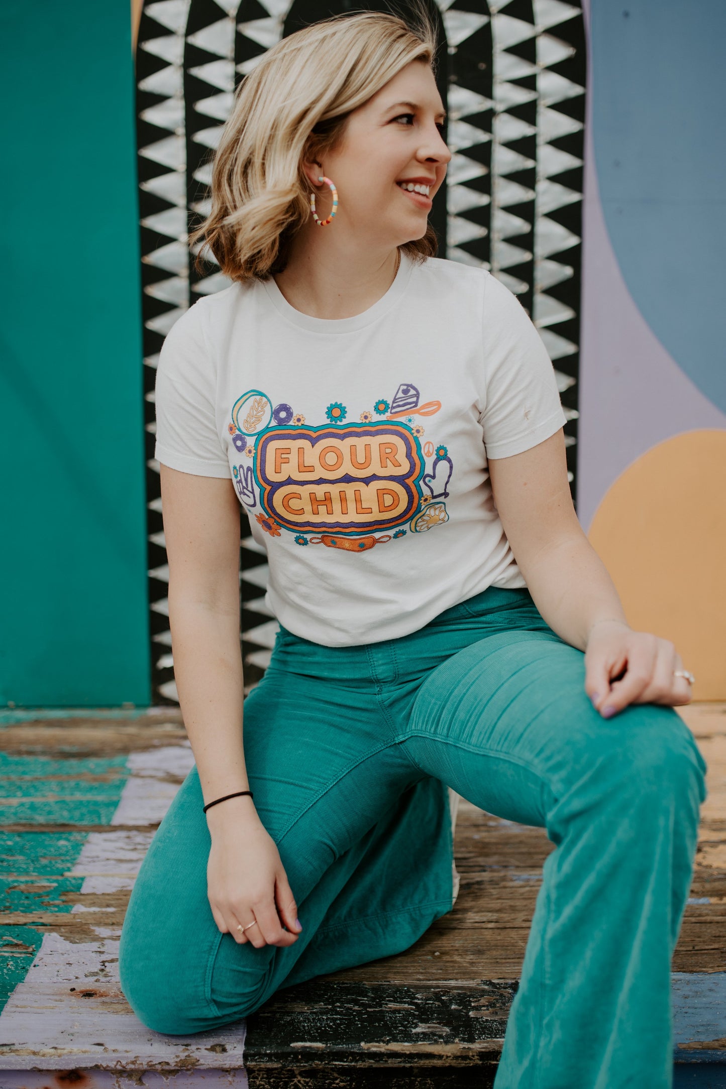 A woman in a white tee with colorful designs and the words Flour Child and teal pants poses in front of a mural