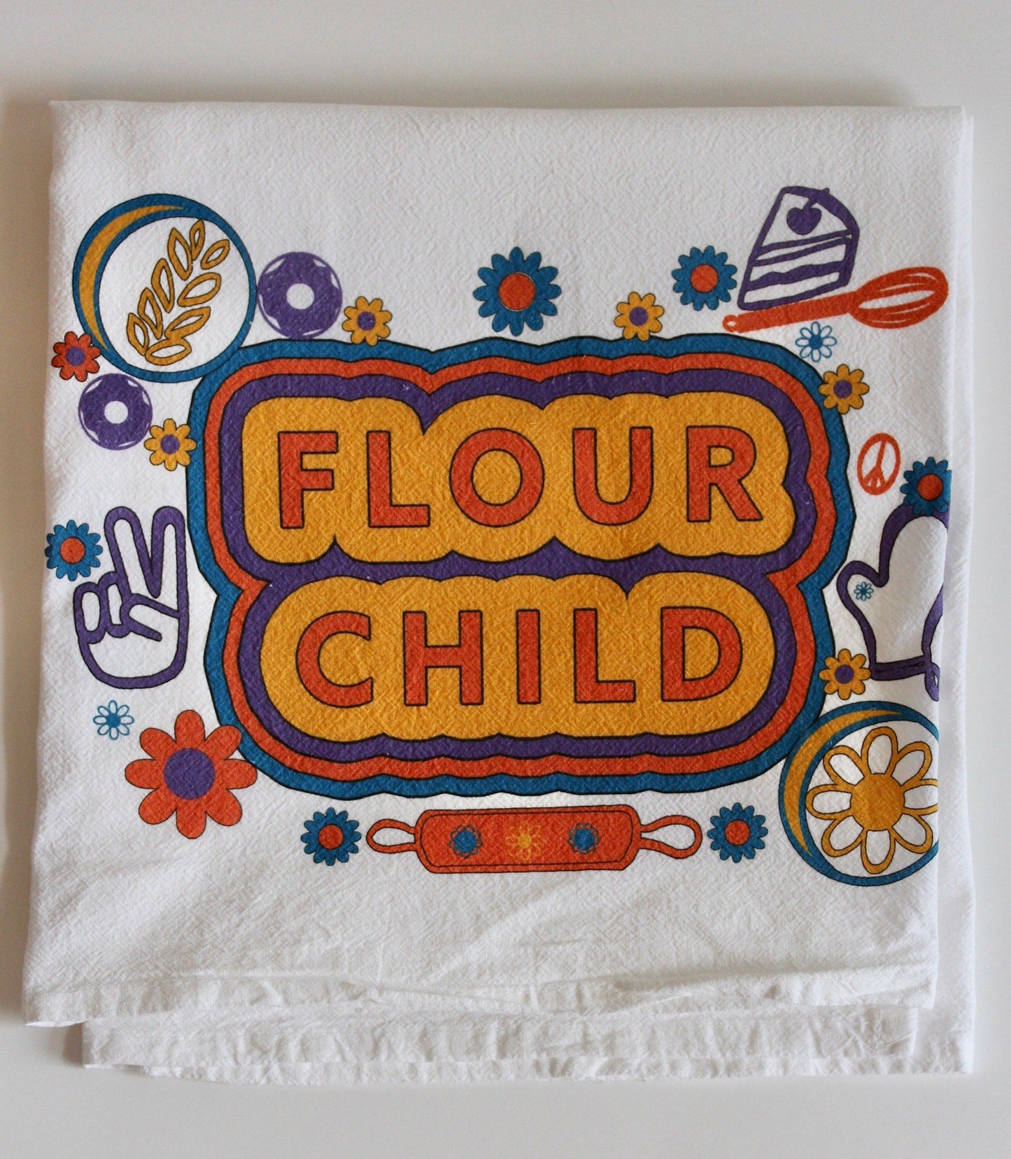 A folded white tea towel with the words Flour Child in block letters and yellow, orange, blue and purple illustrations 