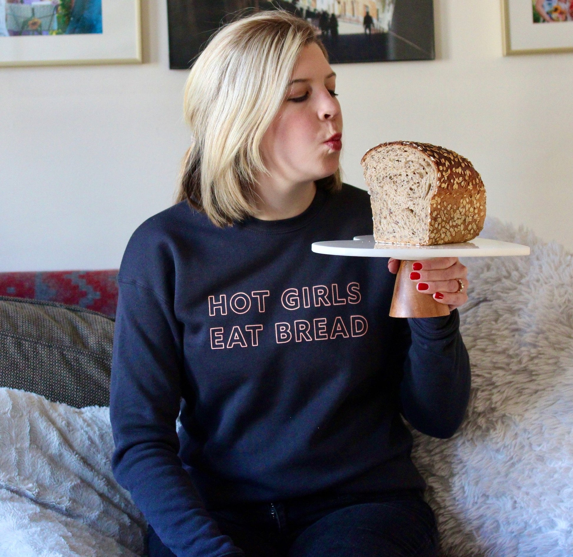 A woman wears a dark grey crewneck that reads "Hot Girls Eat Bread" and holds up a loaf of bread