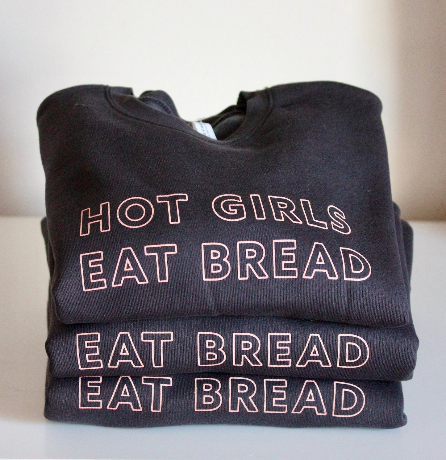 A stack of dark grey sweatshirts with the words Hot Girls Eat Bread in pink block letters