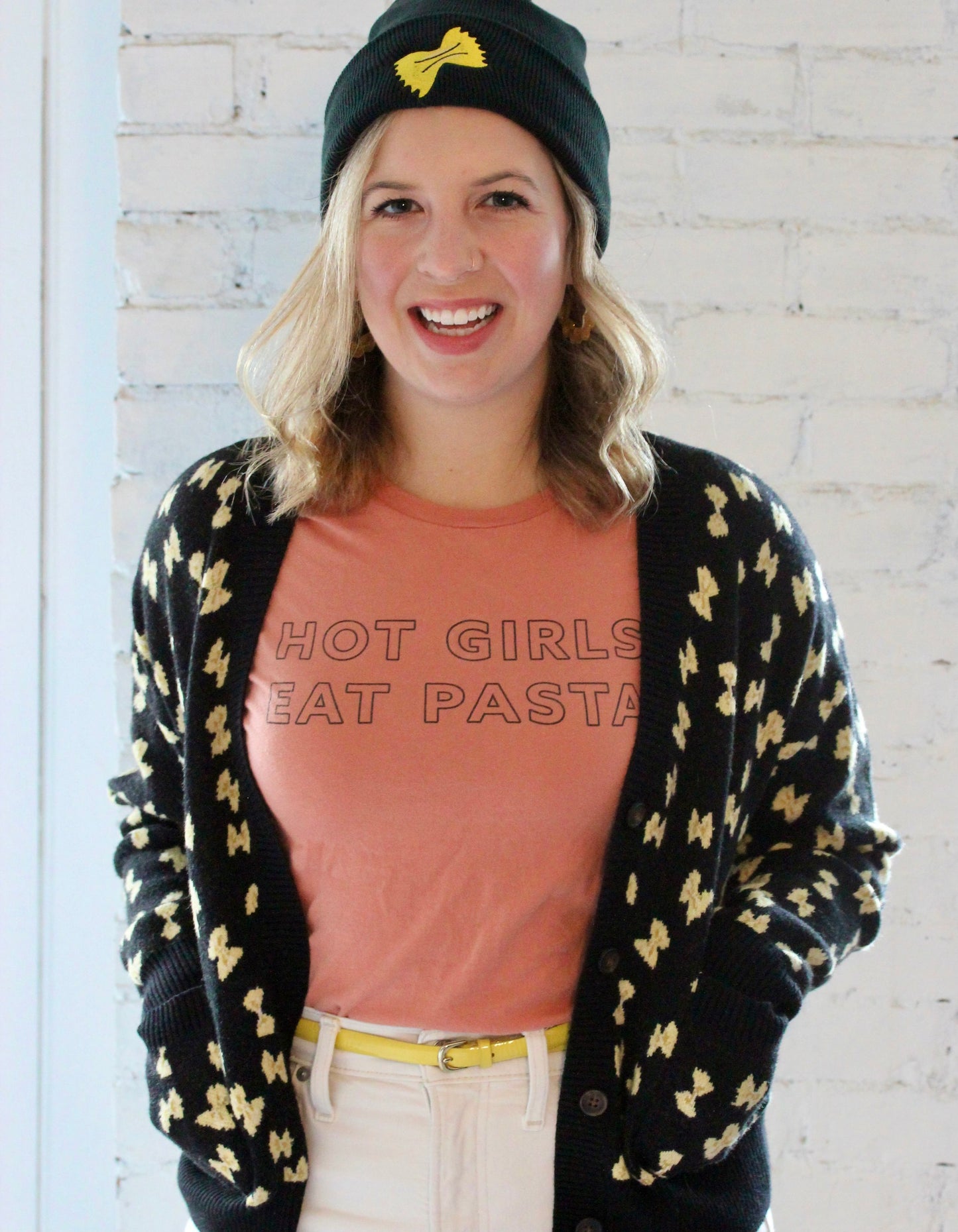 A woman wears a peach tee with the words Hot Girls Eat Pasta and a sweater and hat with a pasta design.