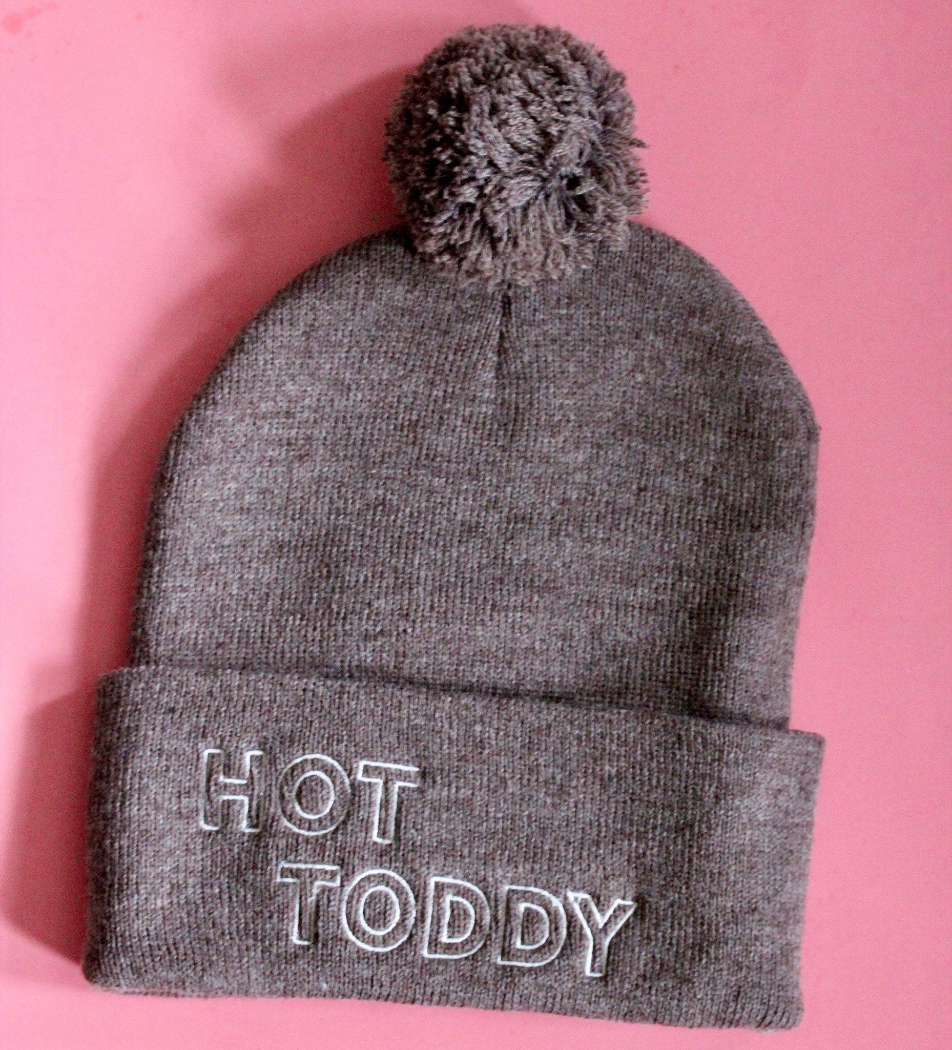 A brown beanie with a pom pom and the words Hot Toddy embroidered in white