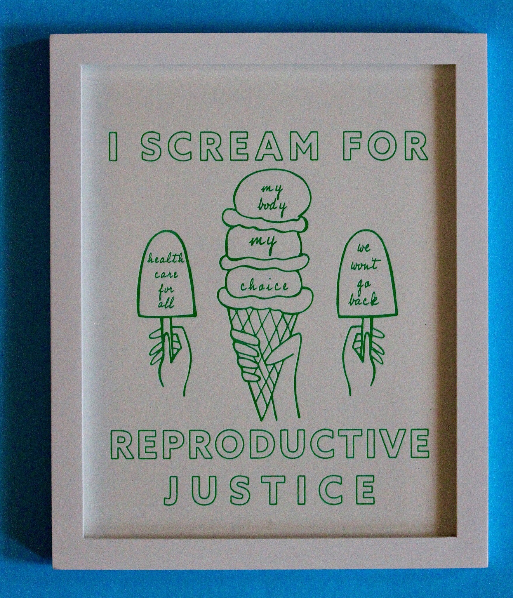A framed print that reads "I Scream for Reproductive Justice" in mint green letters with ice cream illustrations on a blue wall