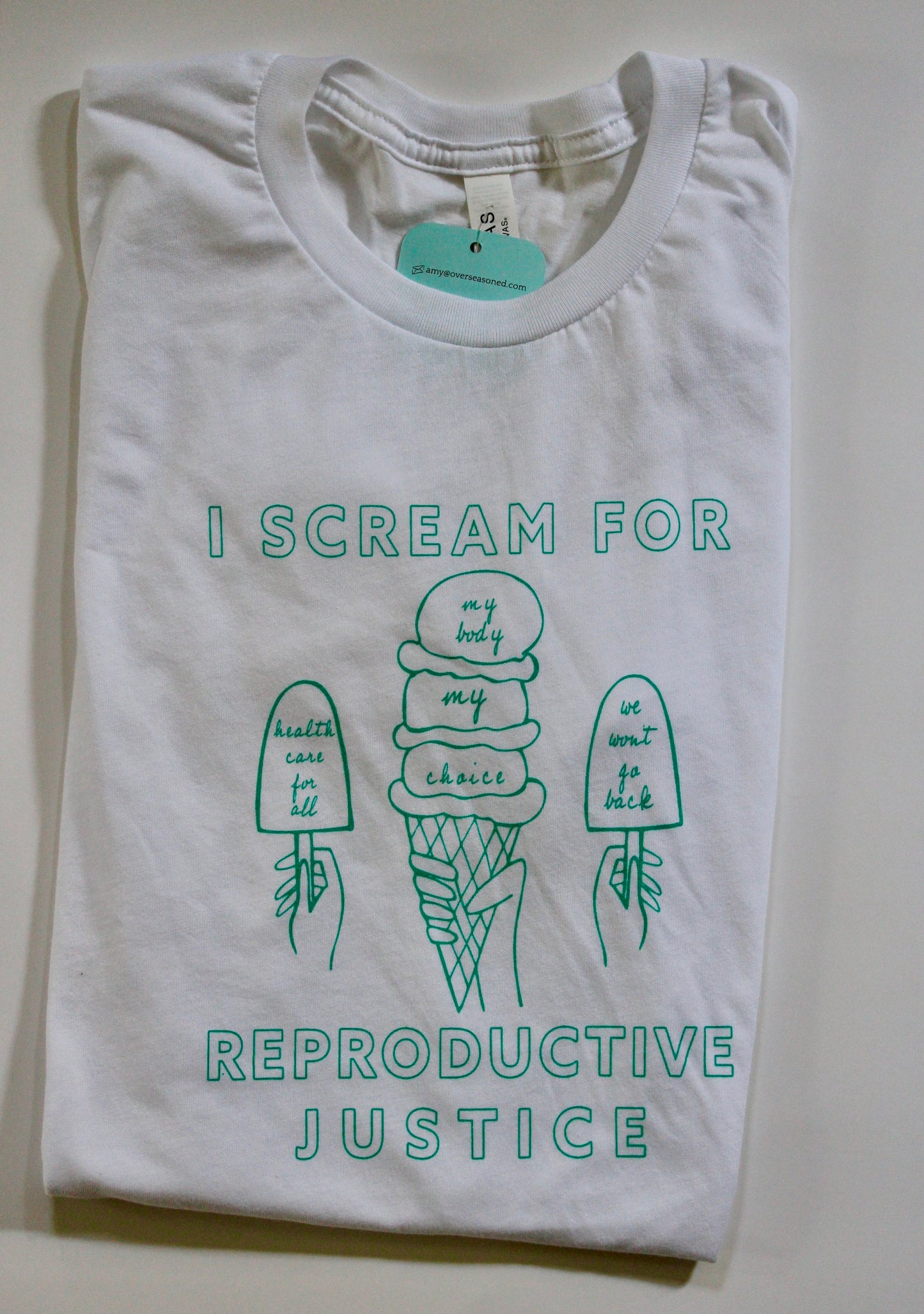 A folded white tee with the words I Scream for Reproductive Justice in mint green with mint green illustrations