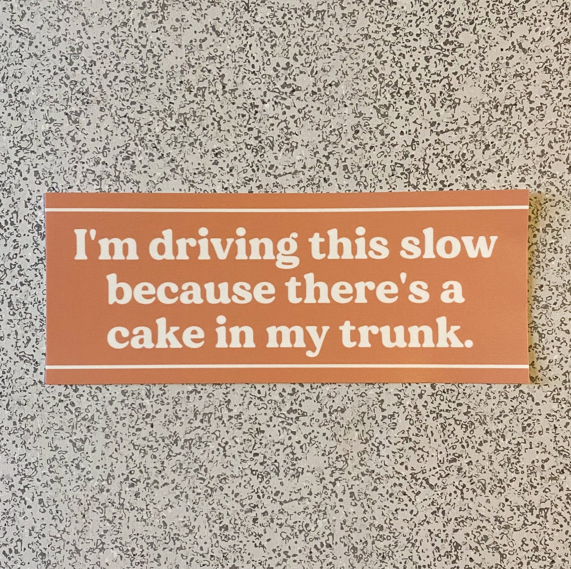 A funny orange bumper sticker with the words "I'm driving this slow because there's a cake in my trunk" in white