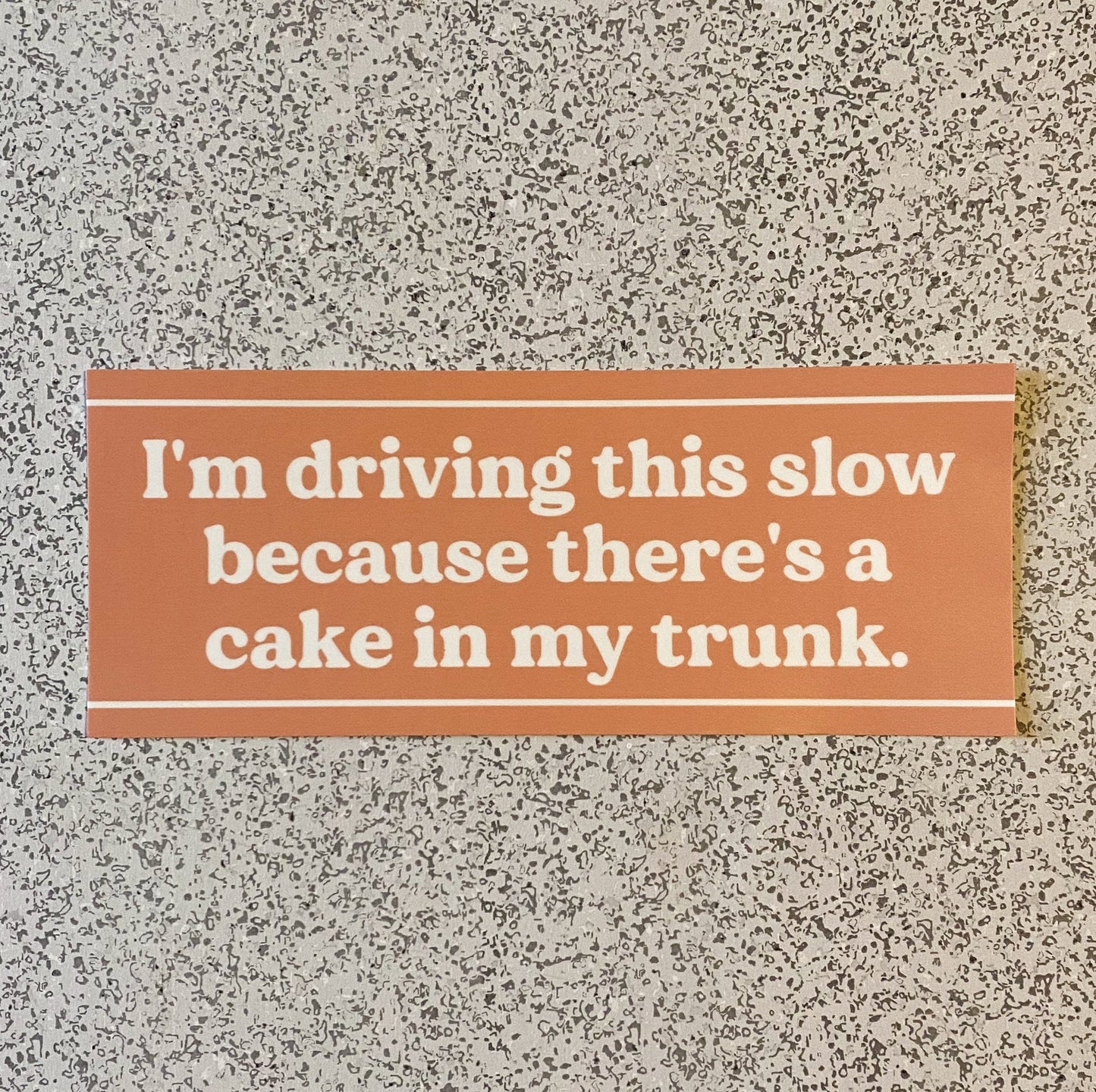 A funny orange bumper sticker with the words "I'm driving this slow because there's a cake in my trunk" in white