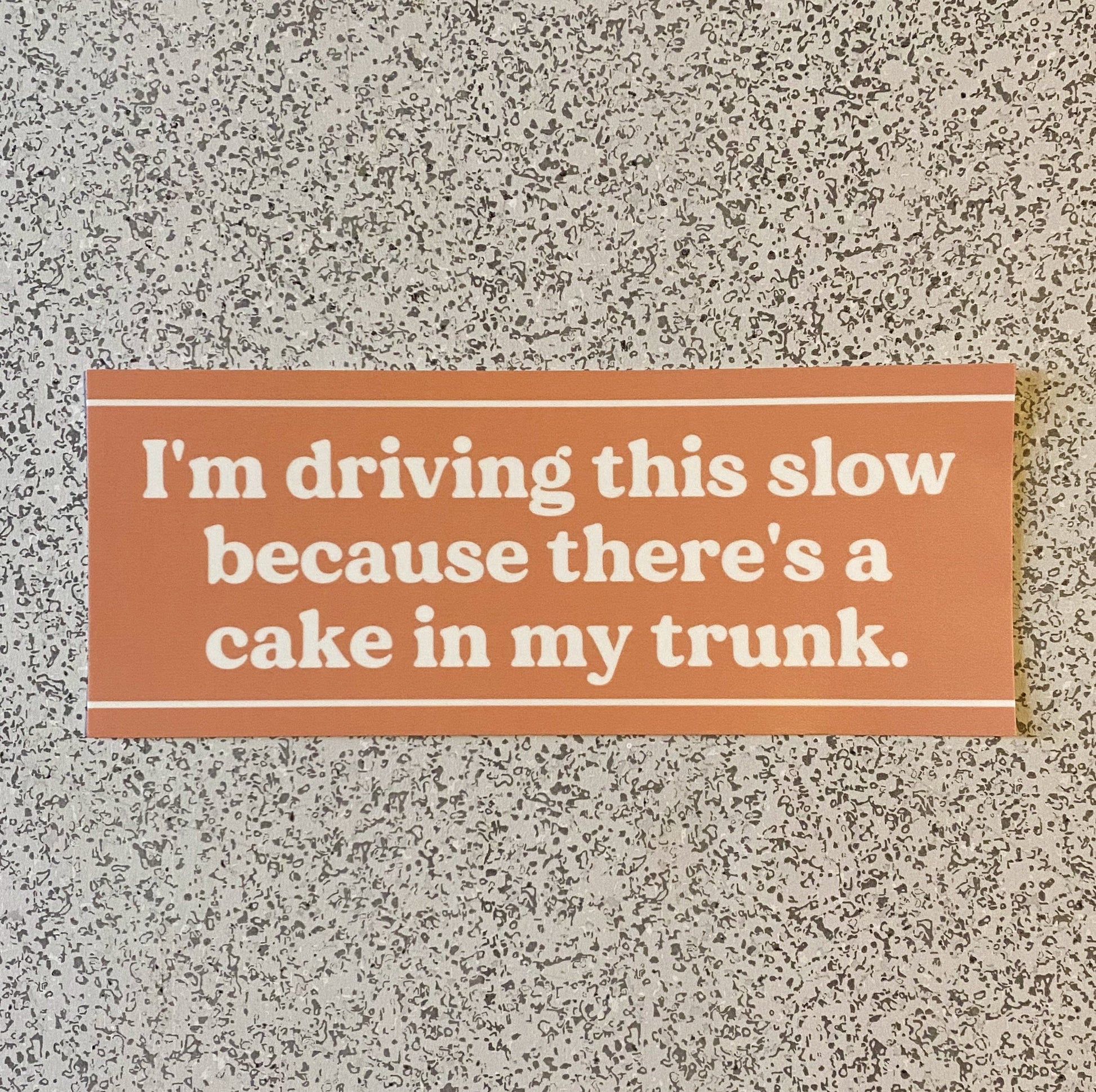 A funny orange bumper sticker with the words "I'm driving this slow because there's a cake in my trunk" in white
