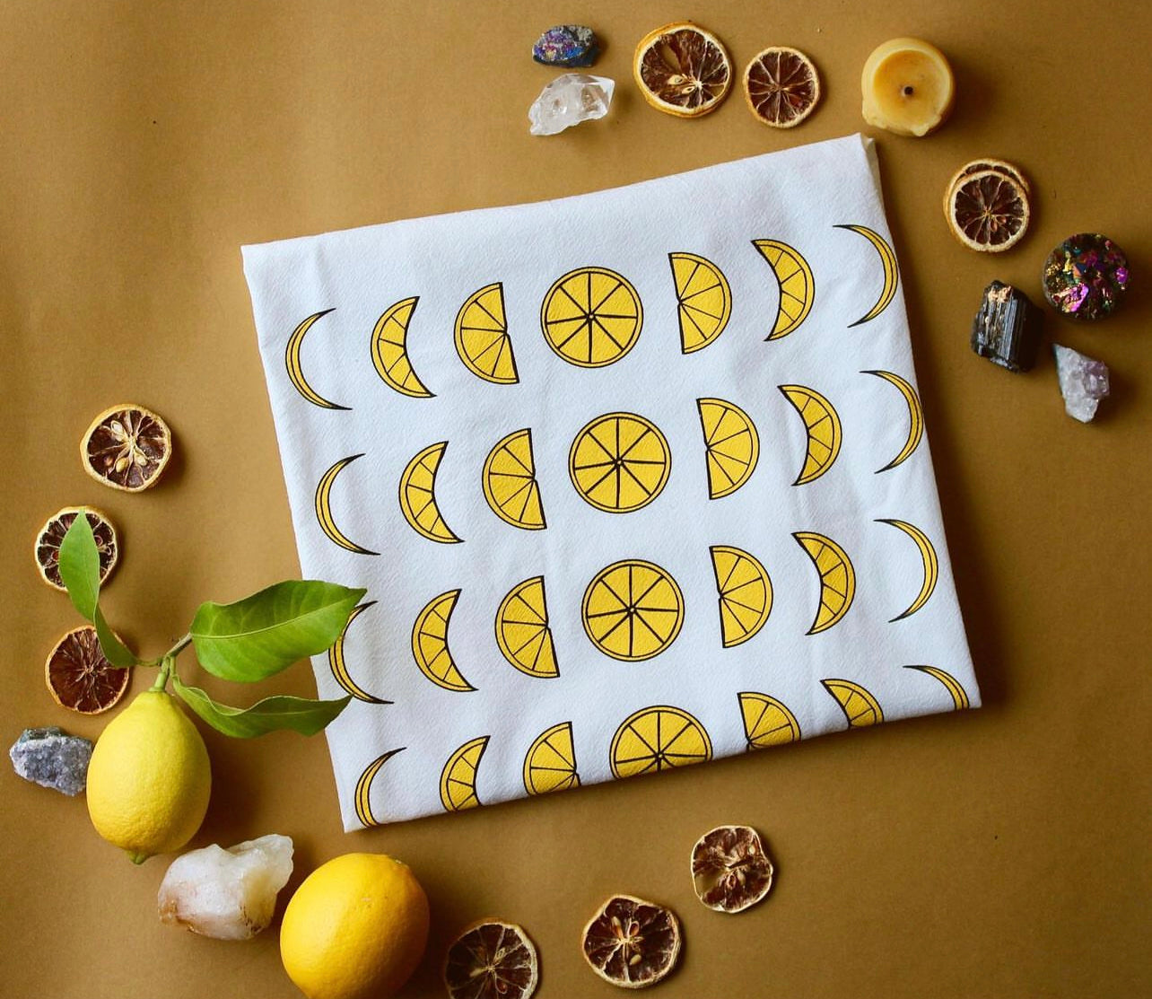 A white tea towel with a yellow lemon moon phase design is surrounded by fresh and preserved lemons