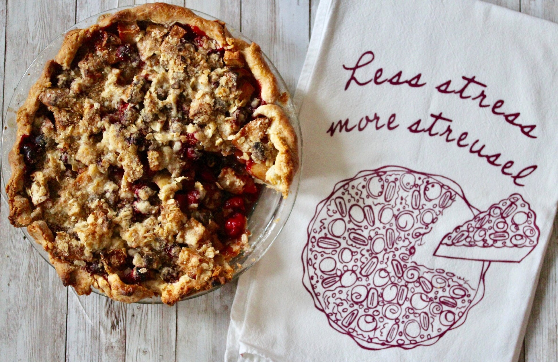 A white tea towel with the words "Less stress, more streusel" and a pie illustration in dark red next to a cranberry peach pie with streusel topping
