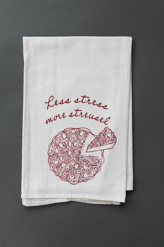 A white tea towel with dark red cursive writing that says "Less stress more streusel" and a pie illustration