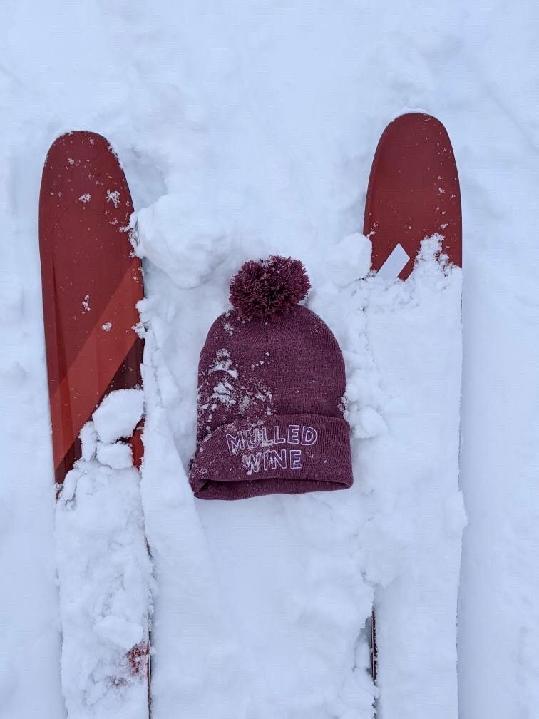 A pink hat with the words "Mulled Wine" in white embroidery in the snow between skis