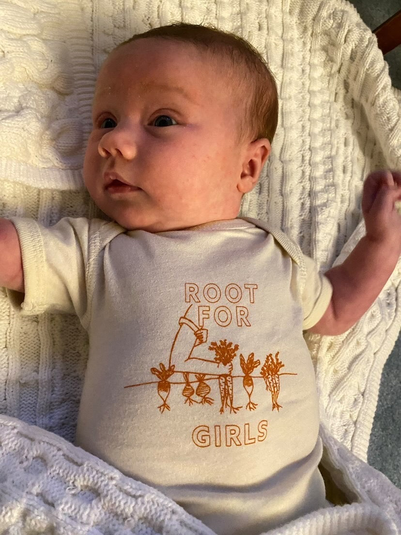 A baby wearing a natural color onesie with the words "Root for Women" in dark orange