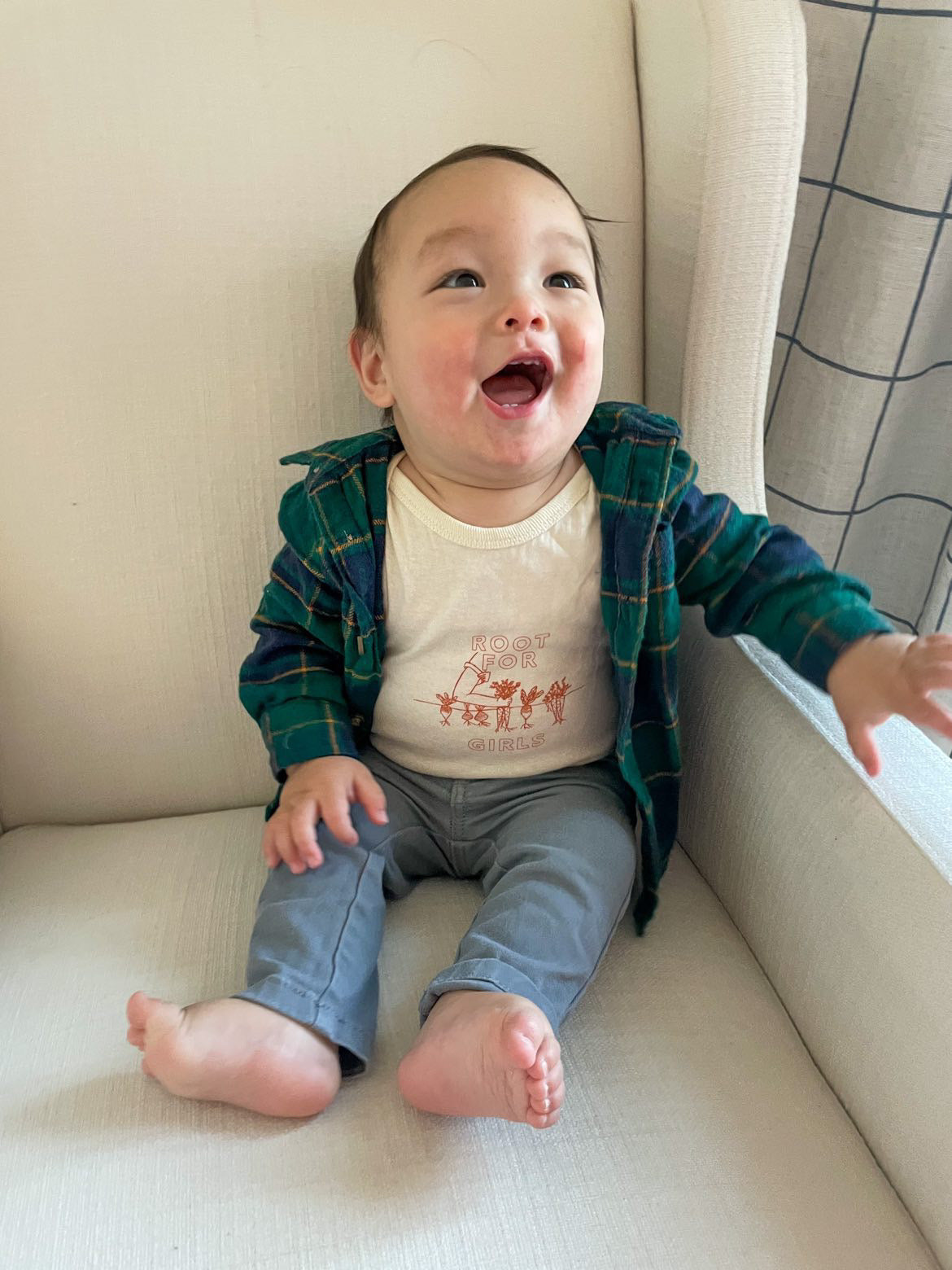 A baby laughs wearing a natural color onesie with the words "Root for Women" and a flannel shirt