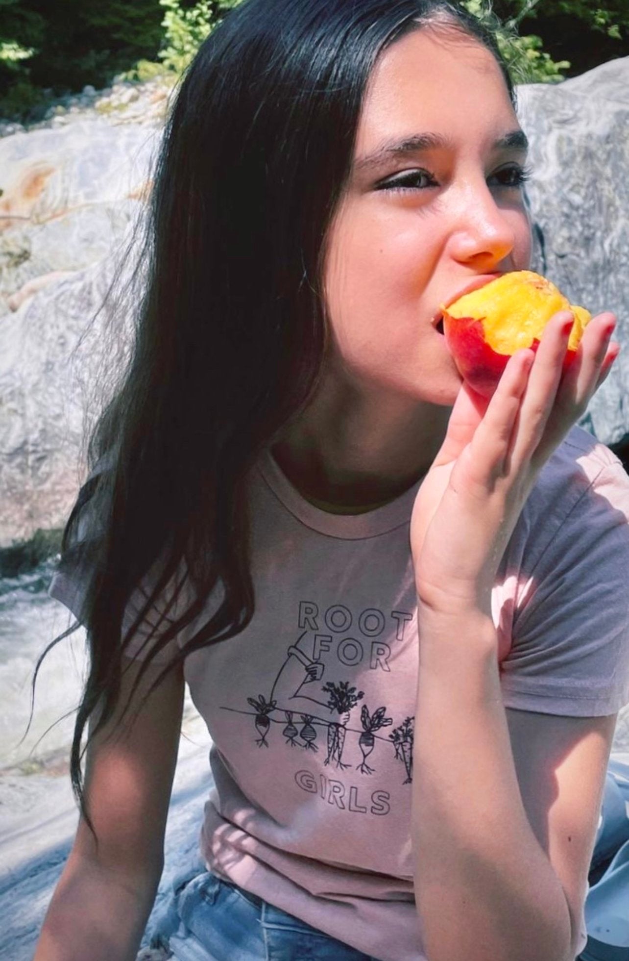 A girl wears a pink tee that reads "Root for Girls" and eats a peach