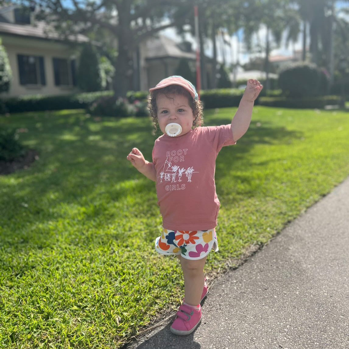 A toddler wears a mauve "Root for Girls" tee with flowered shorts and pink sneakers