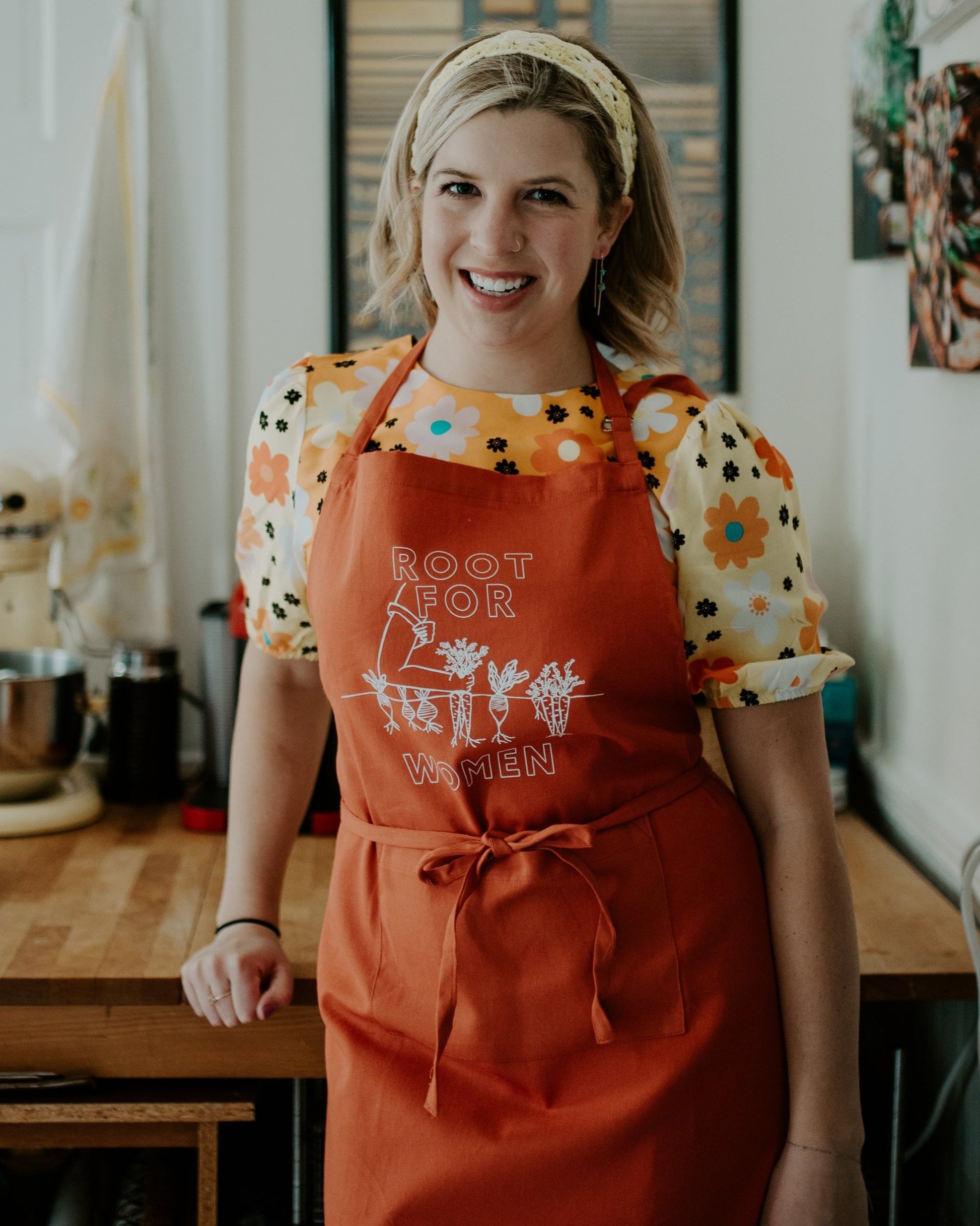 A woman wears an orange "Root for Women" apron over a flowered top