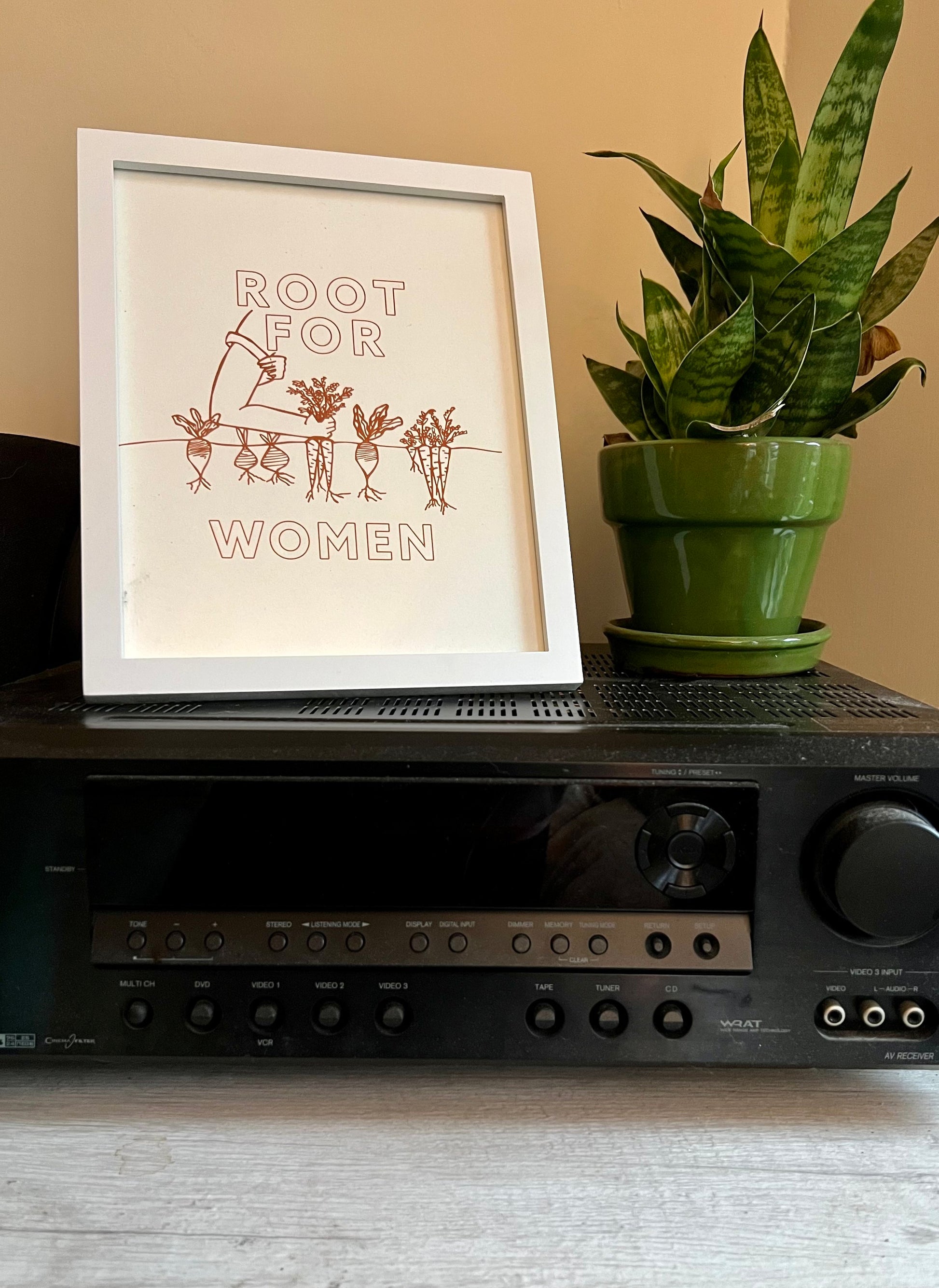 A framed "Root for women" print on a stereo next to a potted plant