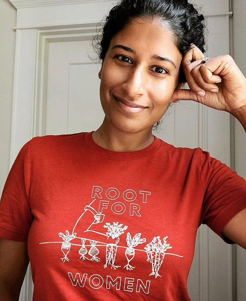 A woman wears a red t-short that reads "Root for Women" with a garden illustration