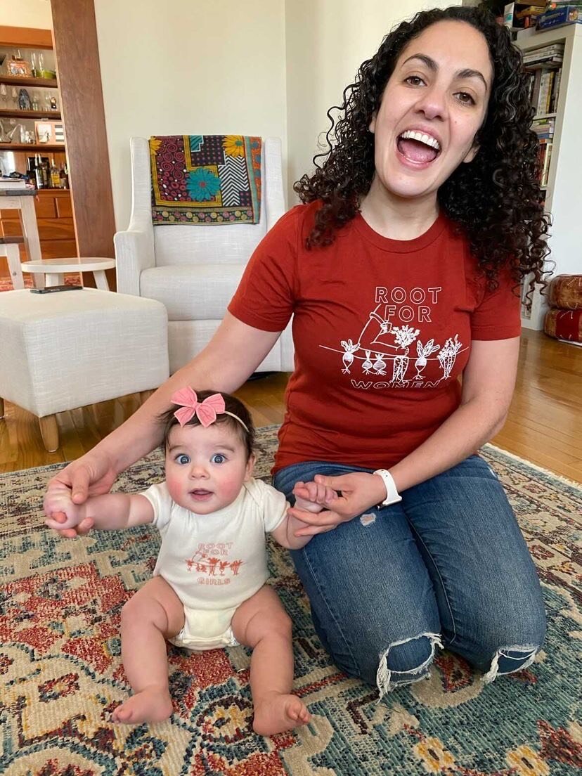 A woman wears a red t-shirt that reads "Root for Women" and holds a baby with a onesie that reads "Root for Girls"