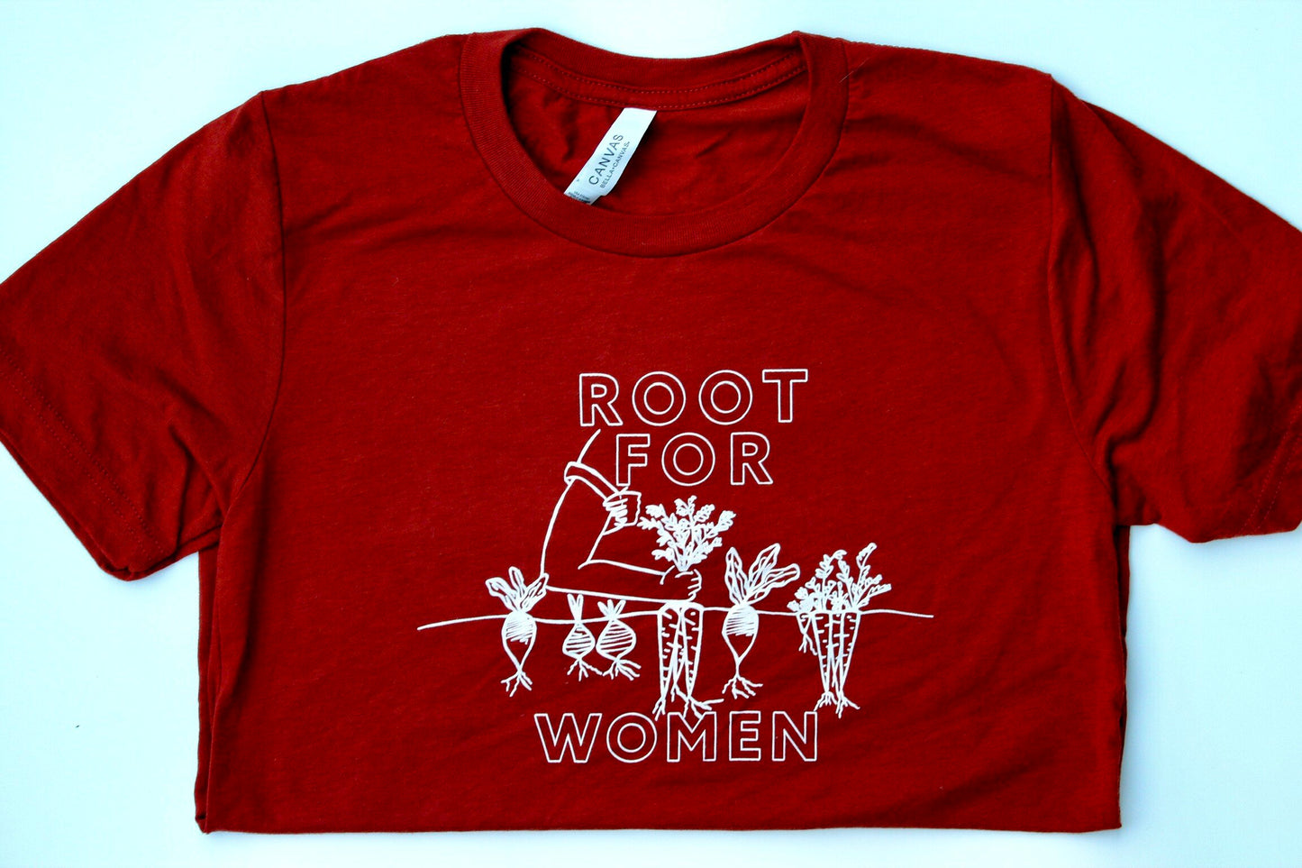 A folded red tee that reads "Root for Women" in block letters with a garden illustration 