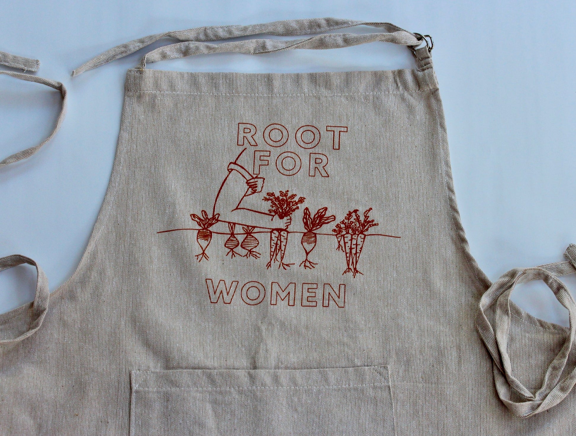 A close up of a natural color chambray apron reads "Root for Women" in carrot orange with a garden illustration
