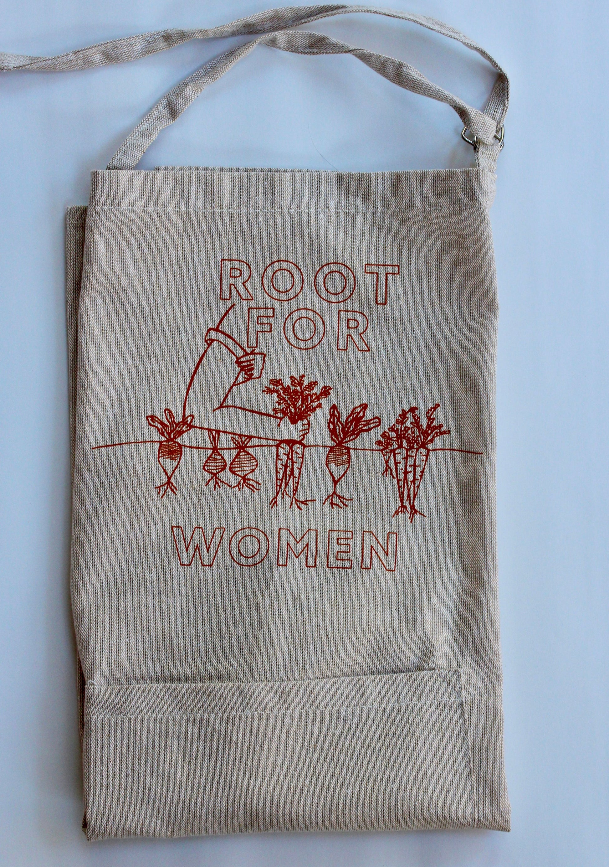 A natural color chambray apron reads "Root for Women" in carrot orange with a garden illustration 