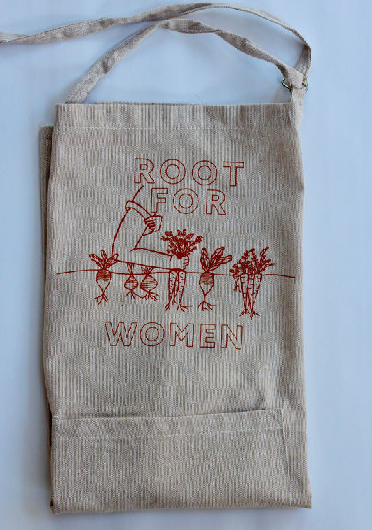 A natural color chambray apron reads "Root for Women" in carrot orange with a garden illustration 