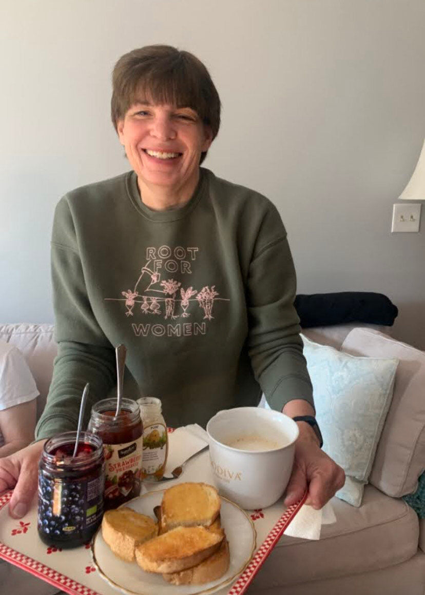 A woman wearing a green "Root for Women" sweatshirt holds a tray of toast and tea
