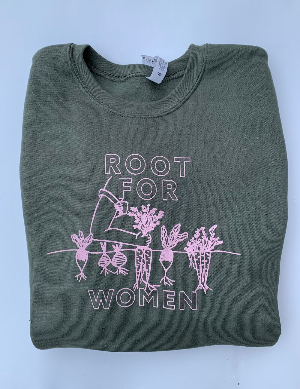 A folded green crewneck reads "Root for Women" in white block letters with a garden illustration