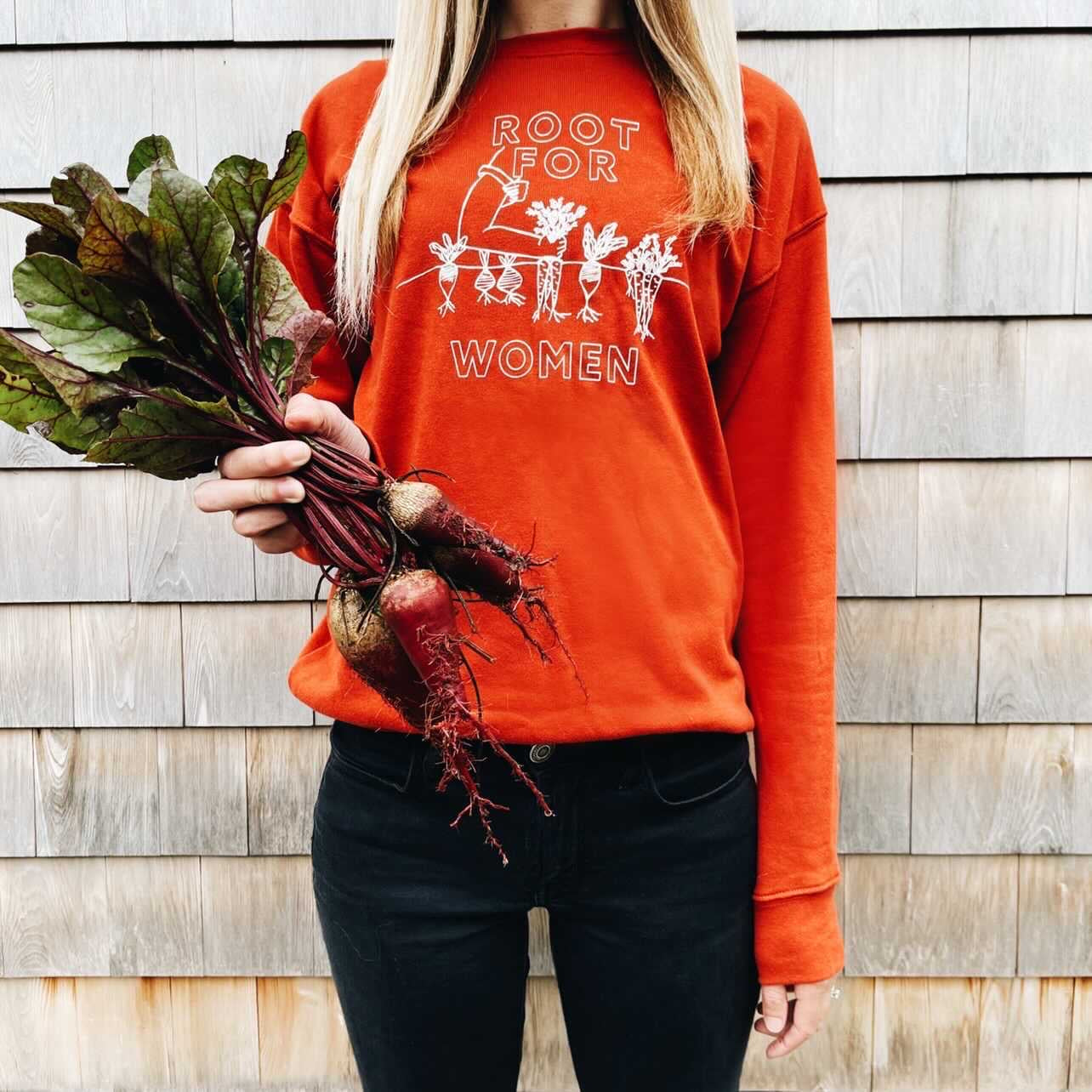 A woman wears a red "Root for Women" crewneck and holds beets