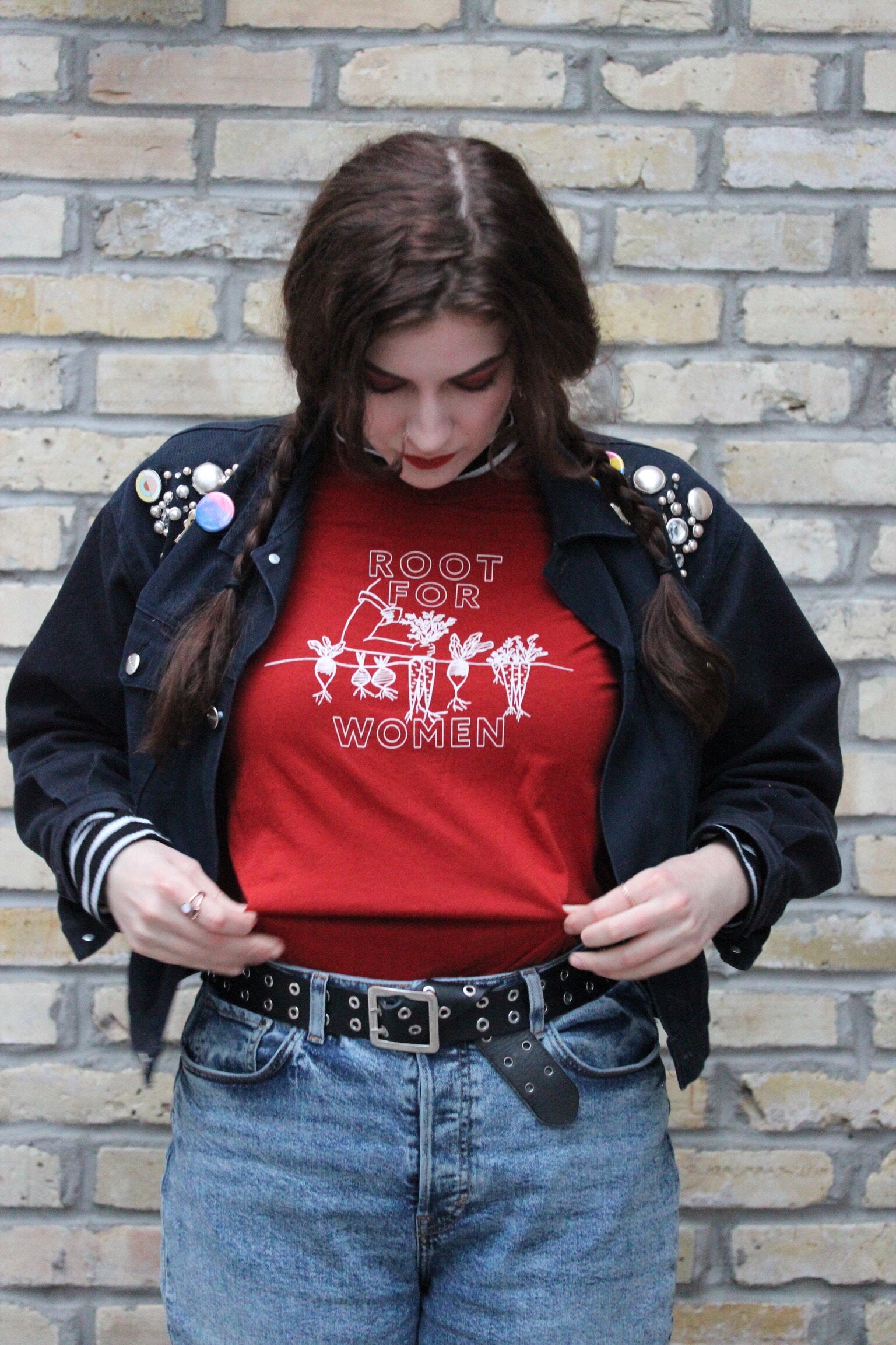 A woman stands in front of a brick wall wearing a red "Root for Women" t-shirt
