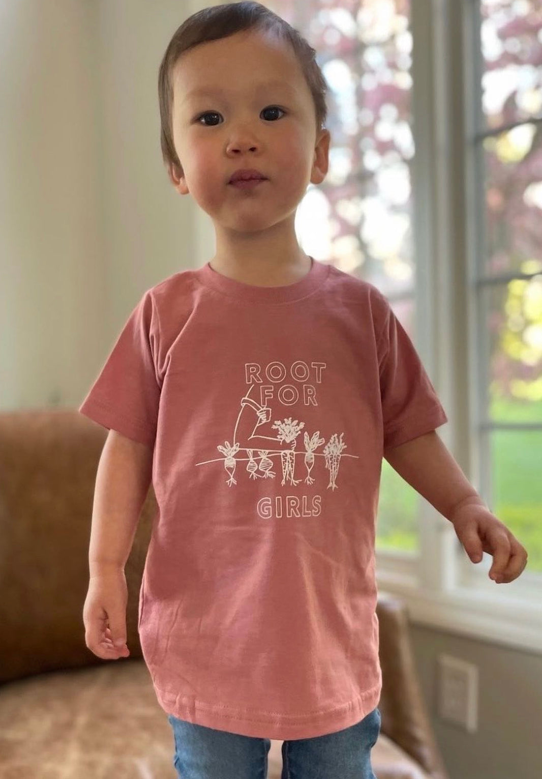 A toddler wears a mauve "Root for Girls" tee with jeans