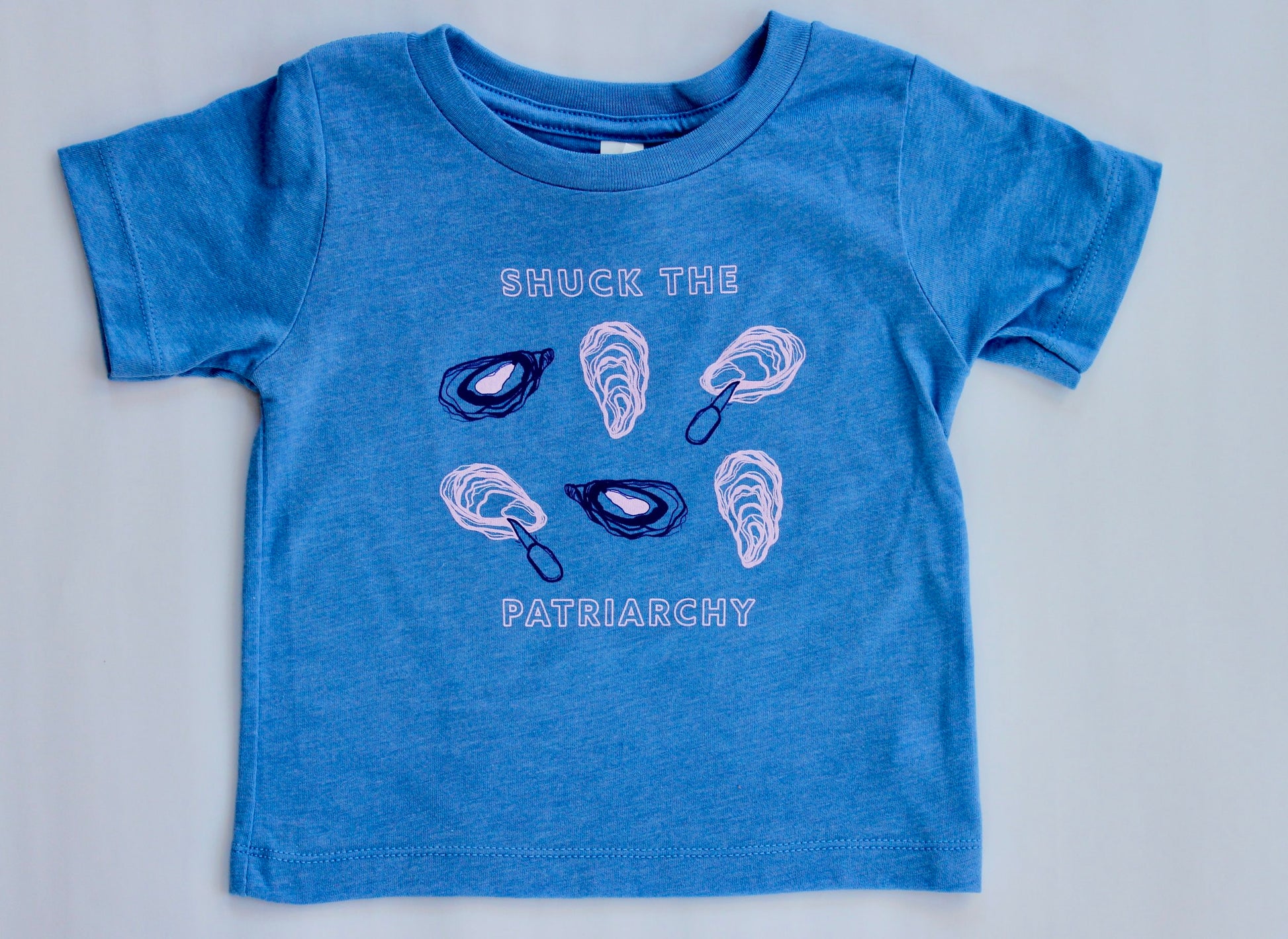 A baby sized tee in bright blue reads "Shuck the Patriarchy" in block letters with oyster illustrations 