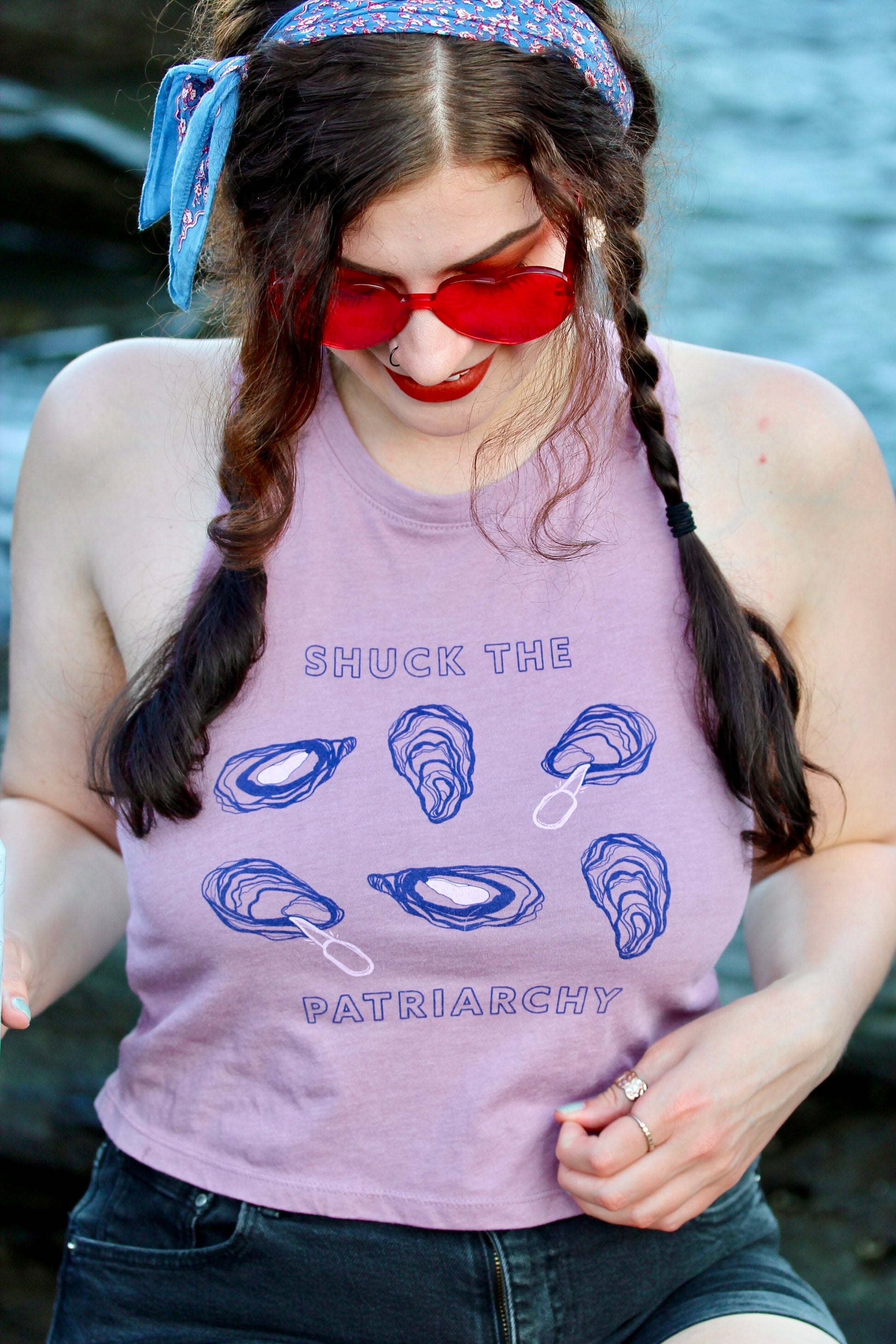 A woman wears heart-shaped sunglasses and a cropped tank that reads "Shuck the Patriarchy" 