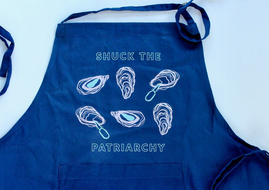 A blue apron with the words "Shuck the Patriarchy" in block letters and oyster illustrations 