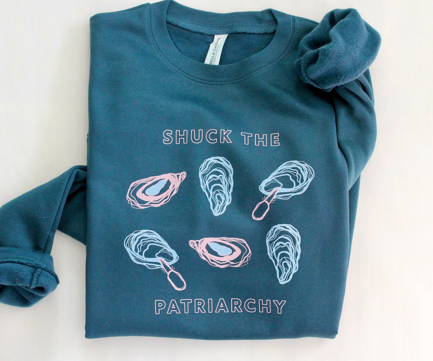 A folded dark teal crewneck sweatshirt reads "Shuck the Patriarchy" with oyster designs