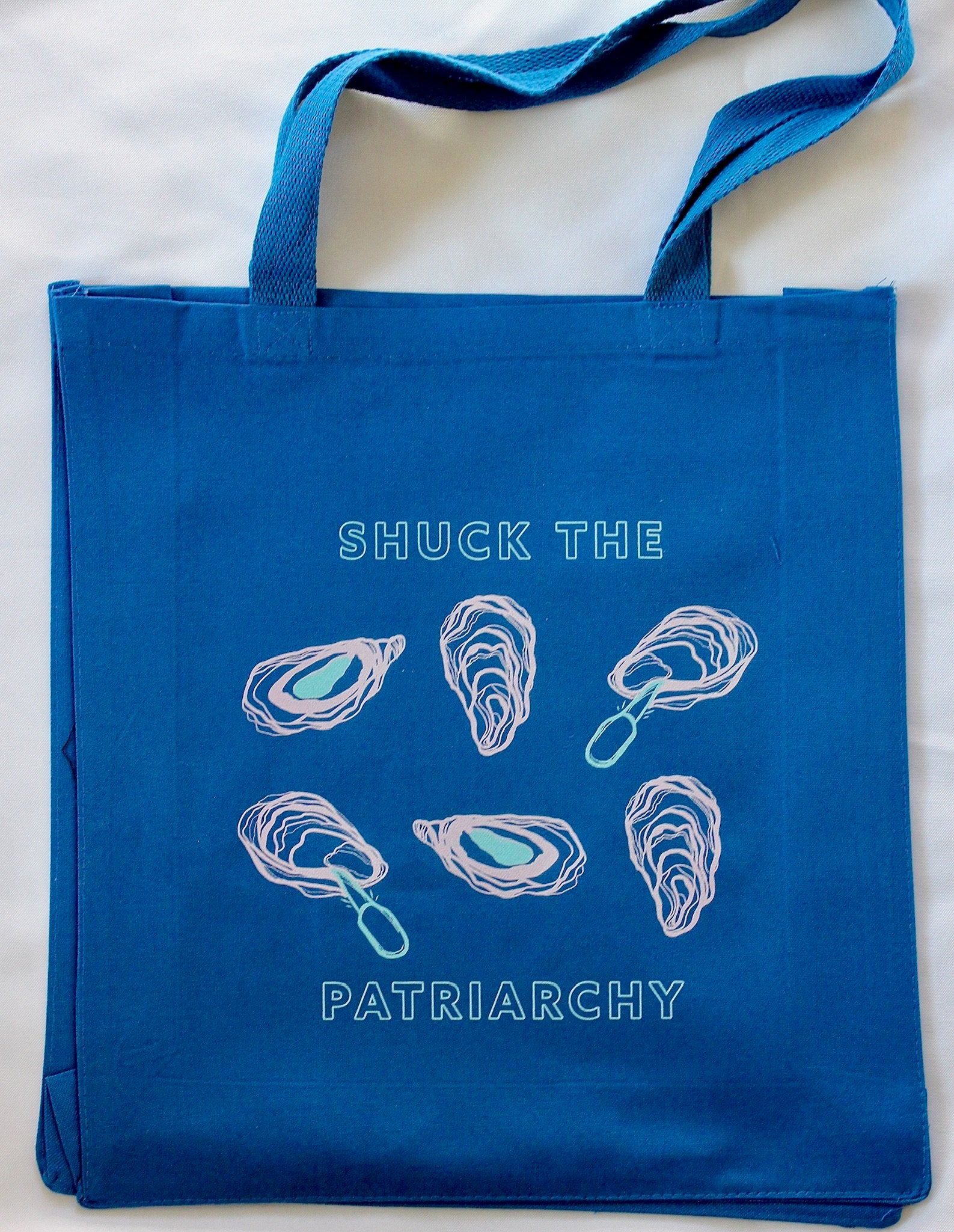 A blue canvas tote reads "Shuck the Patriarchy" in block letters with oyster designs