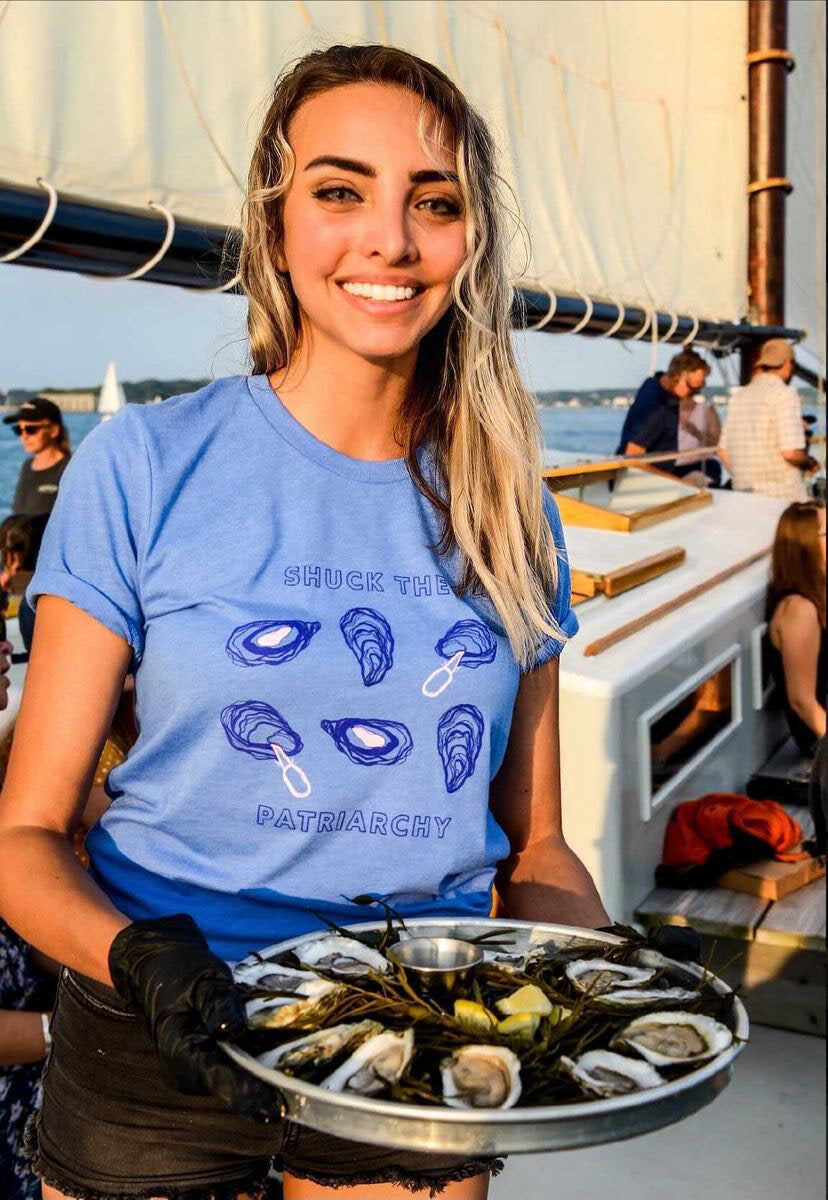 A woman wears a bright blue "Shuck the Patriarchy" t-shirt and holds a plate of oysters on a sailboat