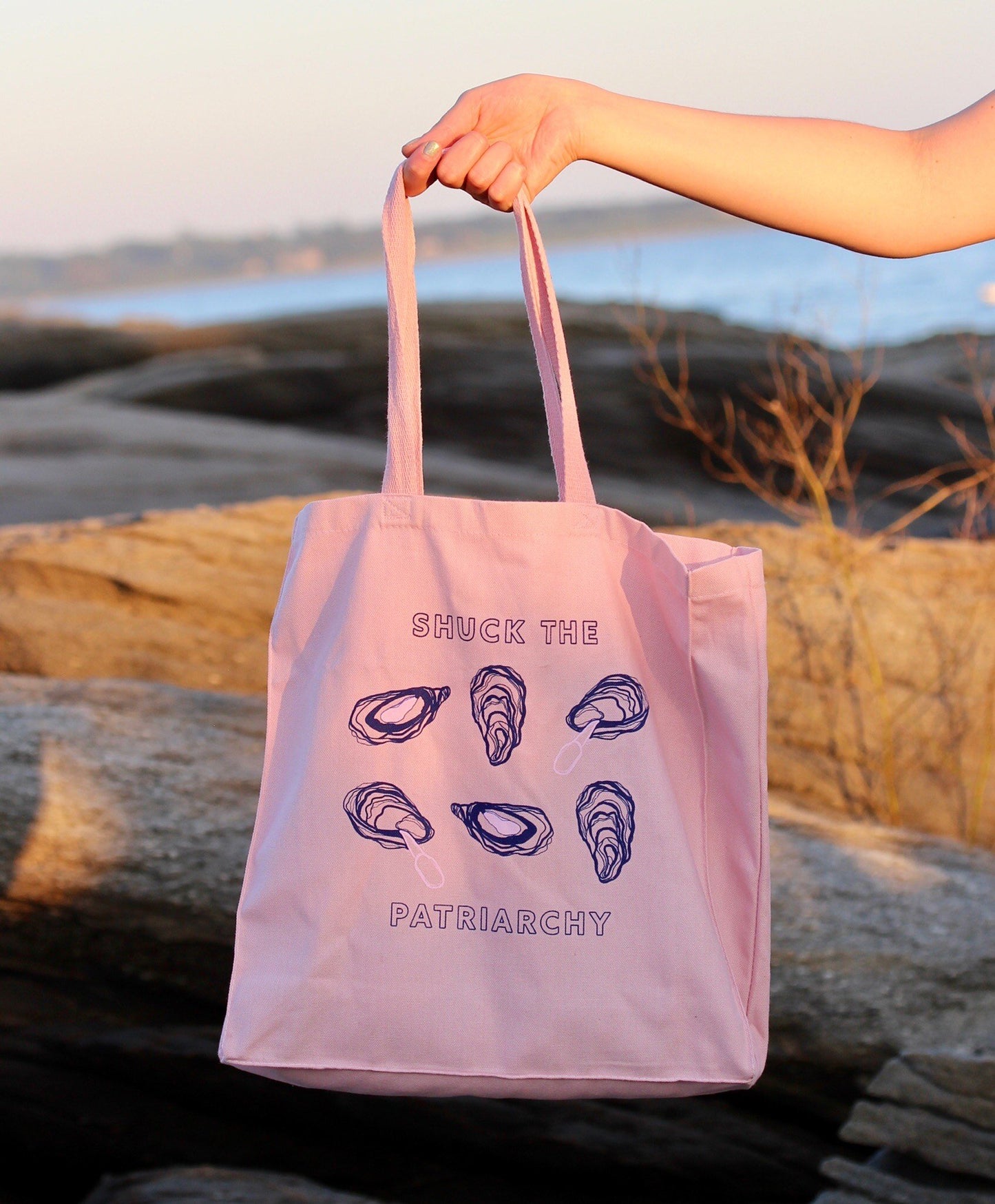 A woman holds a pink tote that reads "Shuck the Patriarchy" in block letters