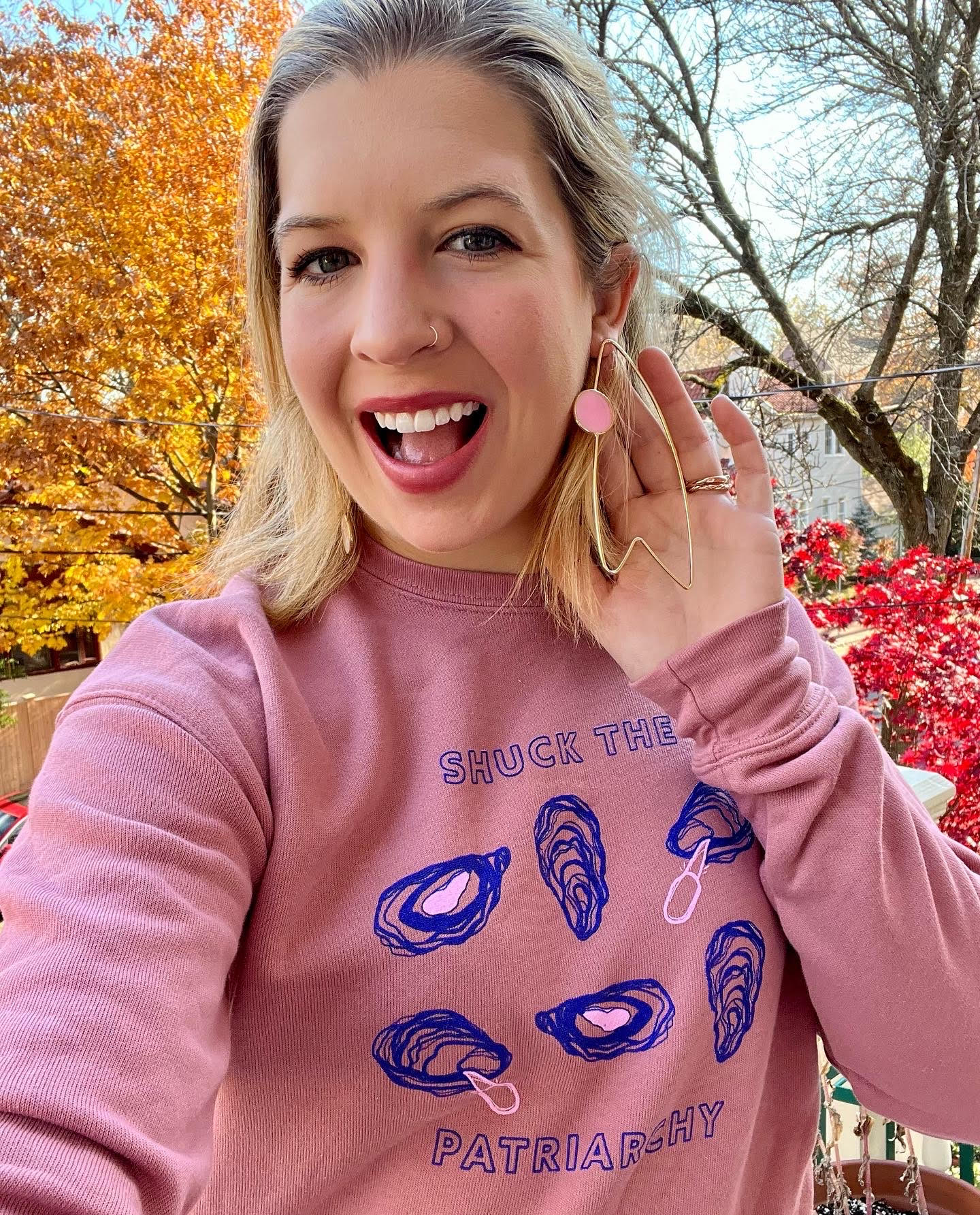 A woman wears a pink "Shuck the Patriarchy" crewneck with matching earrings