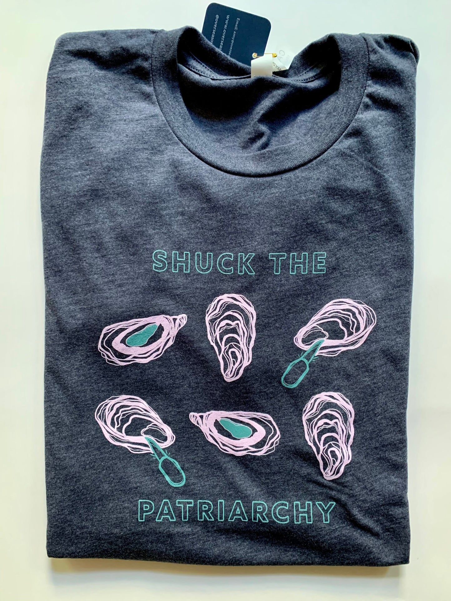 A folded heather navy tee with "Shuck the Patriarchy" in block letters and oyster designs