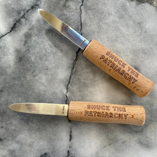 A pair of oyster shucking knives with a wooden handle that reads "Shuck the Patriarchy" in block letters