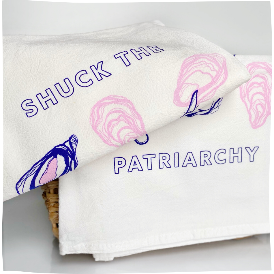 Two folded white tea towels read "Shuck the Patriarchy" in block letters