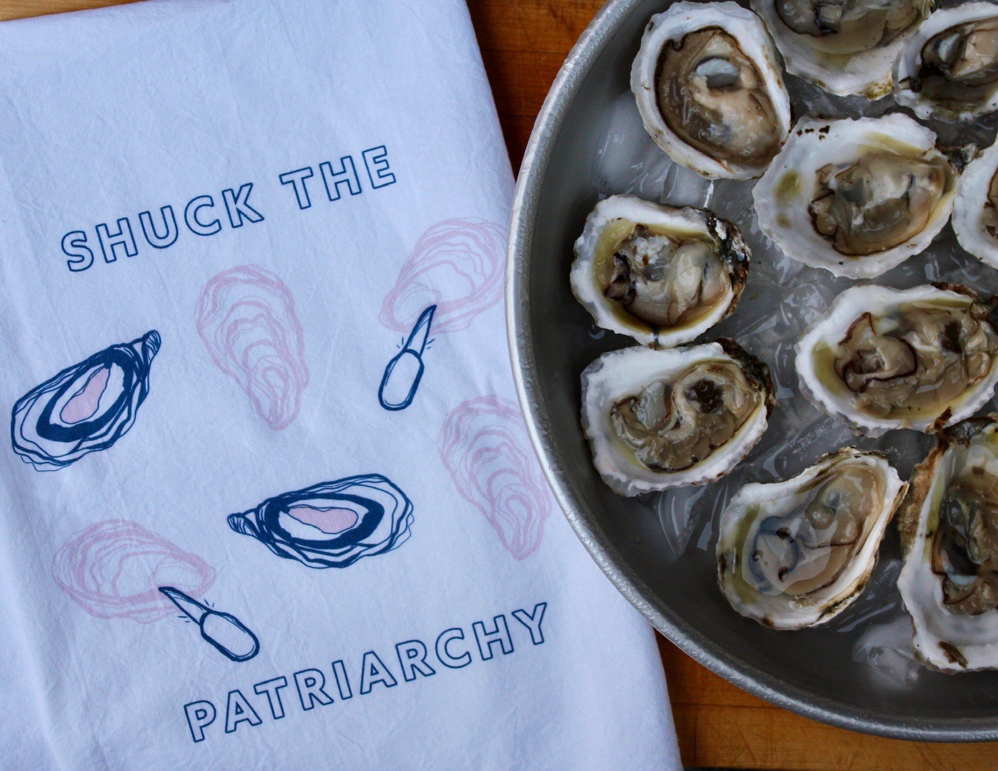A white tea towel that reads "Shuck the Patriarchy" sits next to a platter of oysters on ice