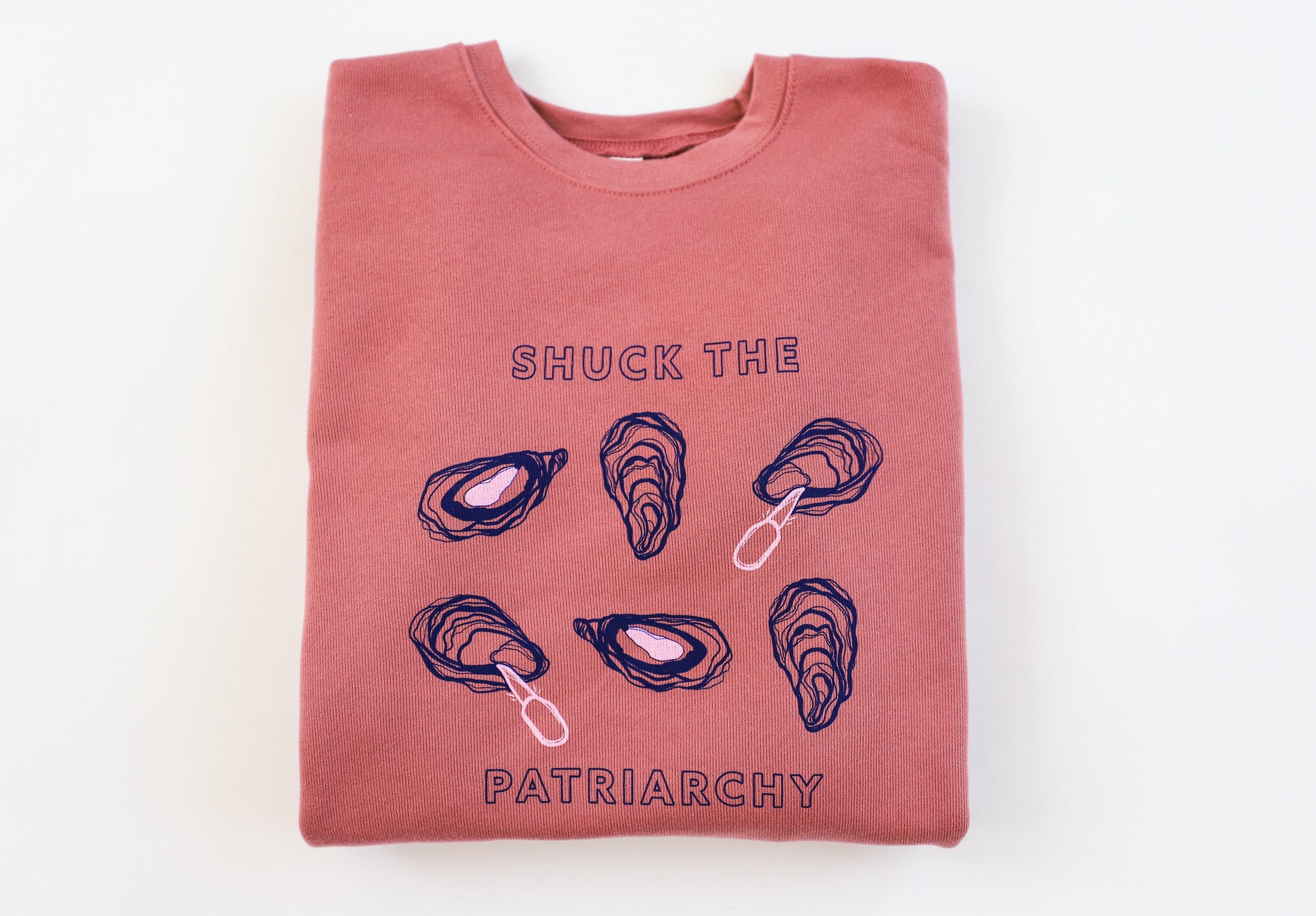 A folded pink crewneck reads "Shuck the Patriarchy" in blue block letters with oyster designs