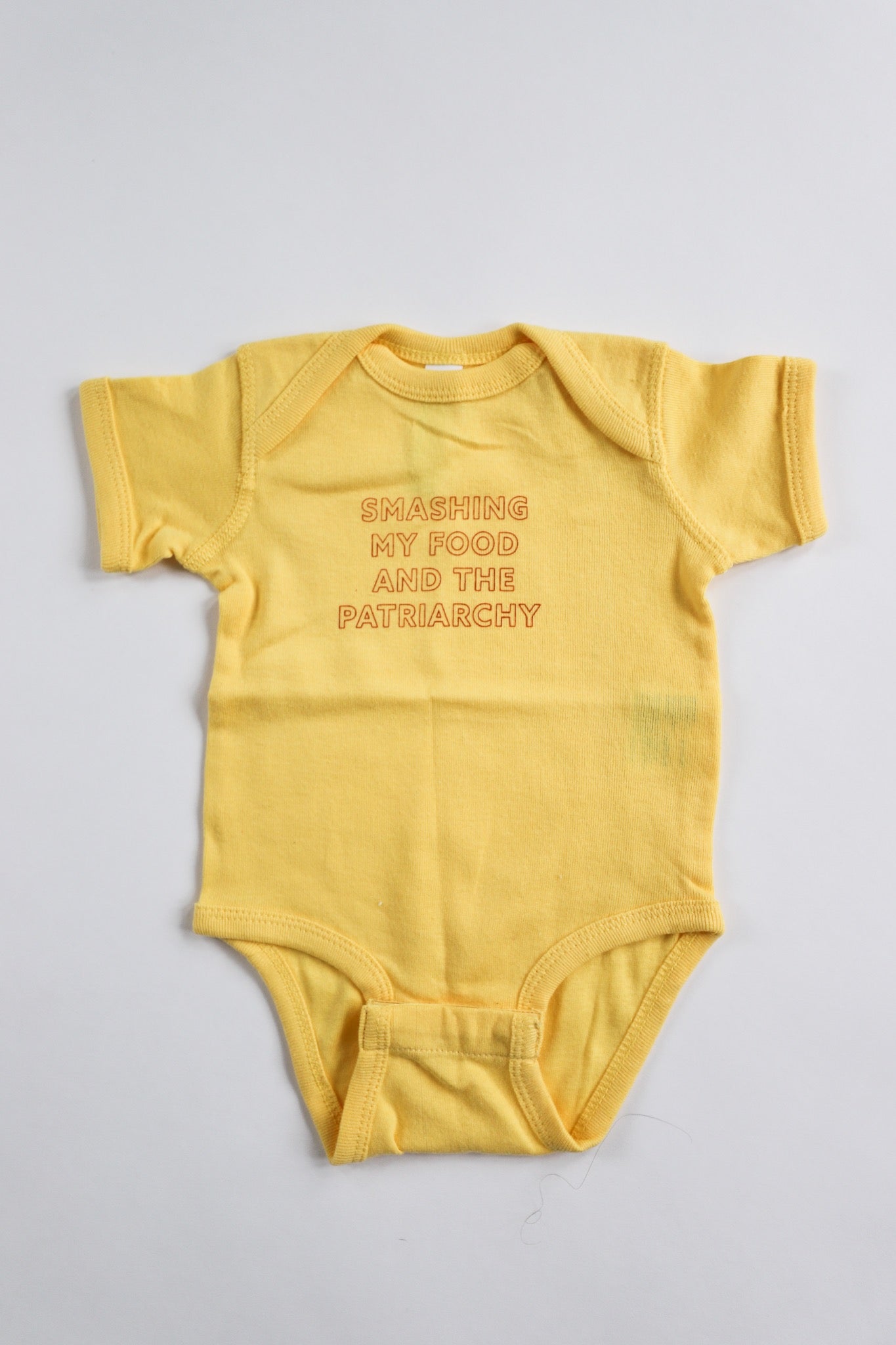 A yellow baby onesie reads "Smashing my Food and the Patriarchy" in red block letters